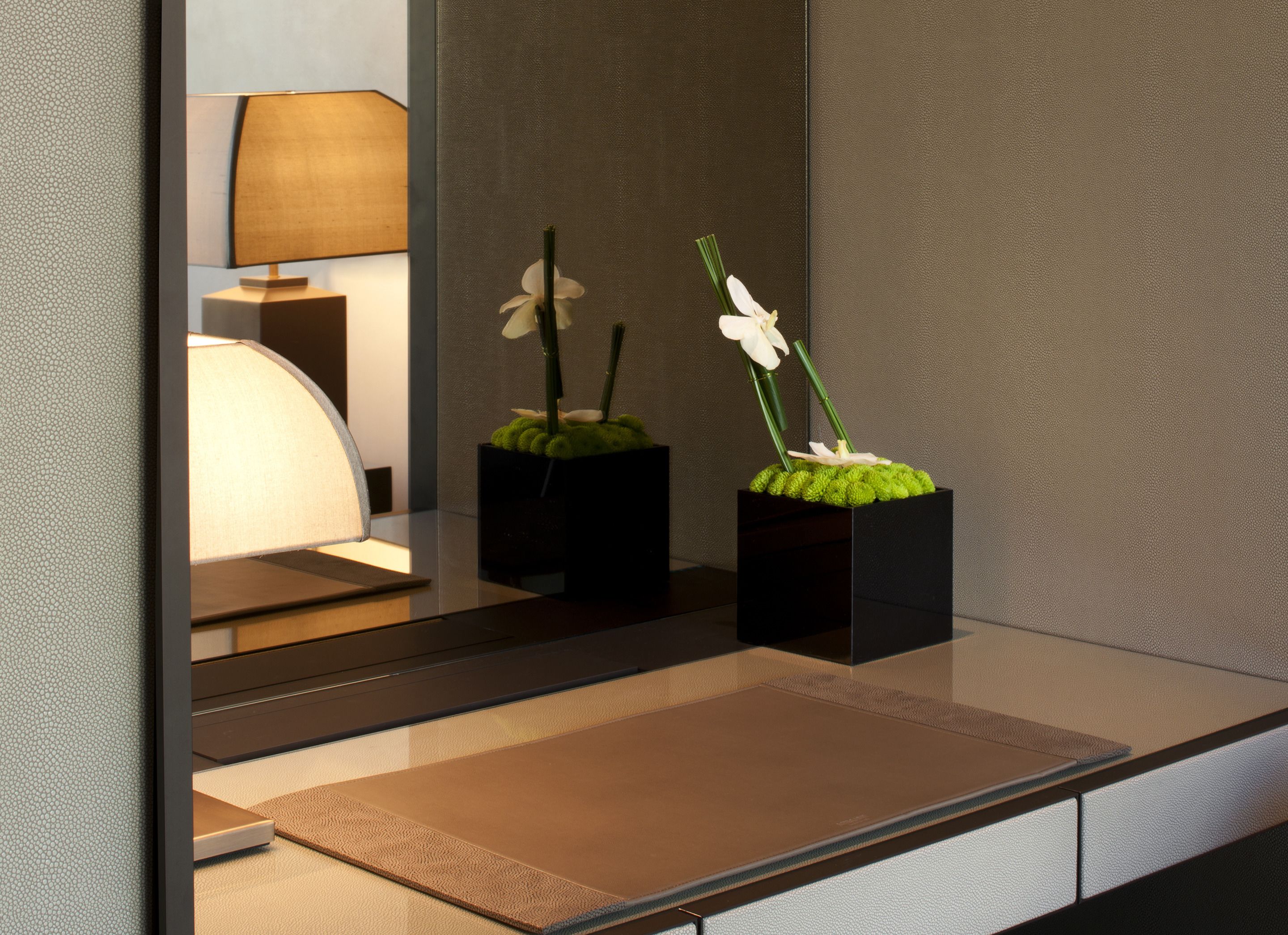 Un interno moderno con una superficie riflettente, due vasi quadrati neri con muschio verde e fiori bianchi, e una lampada sullo sfondo.