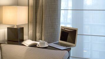 Ein modernes Hotelzimmer mit einem Laptop auf einem Nachttisch, einer Lampe und Blick ins Freie durch große Fenster.