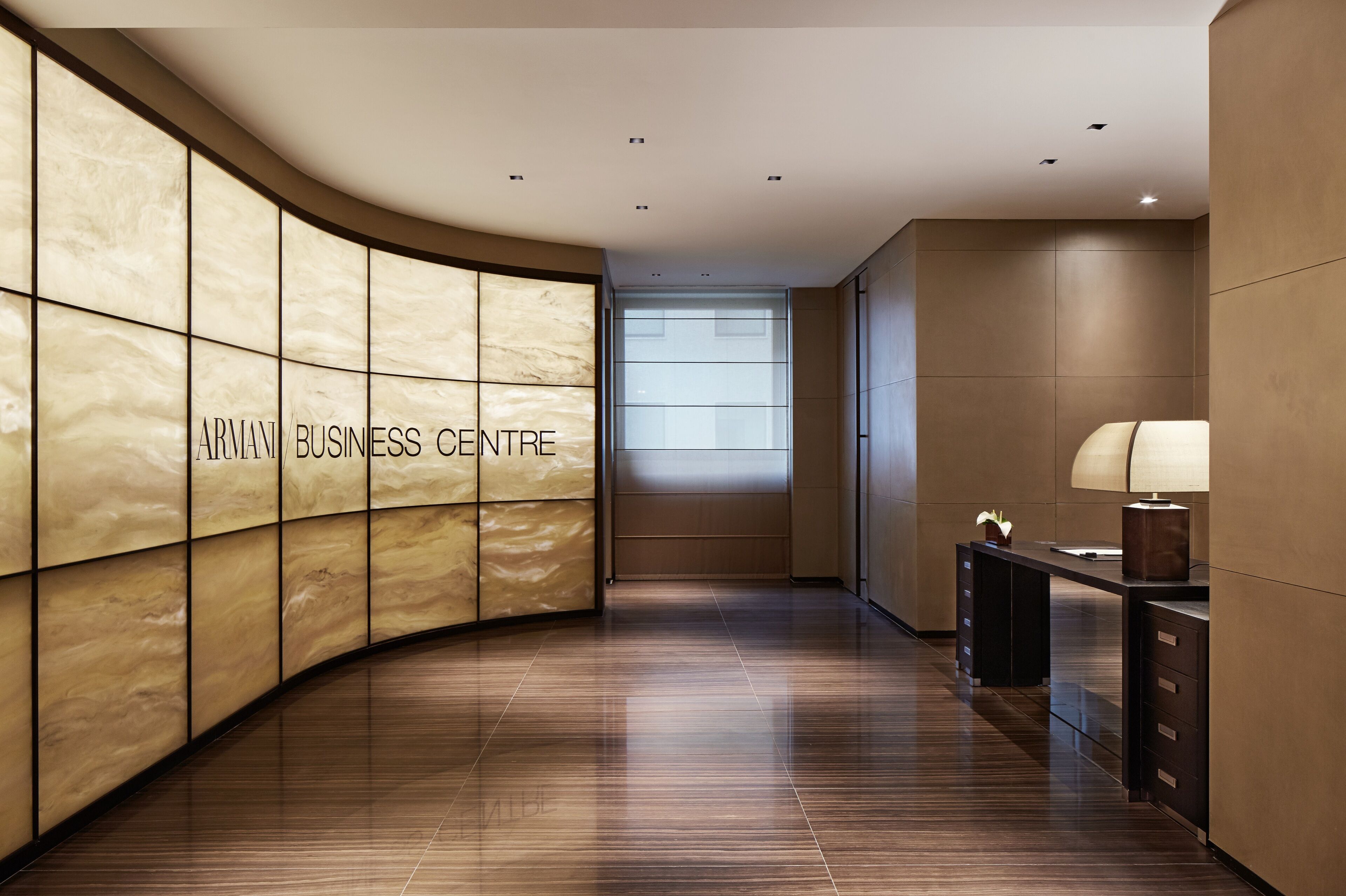 Un interior moderno con una pared curva que muestra 'ARMANI BUSINESS CENTRE' y una zona de recepción con escritorio y lámpara.