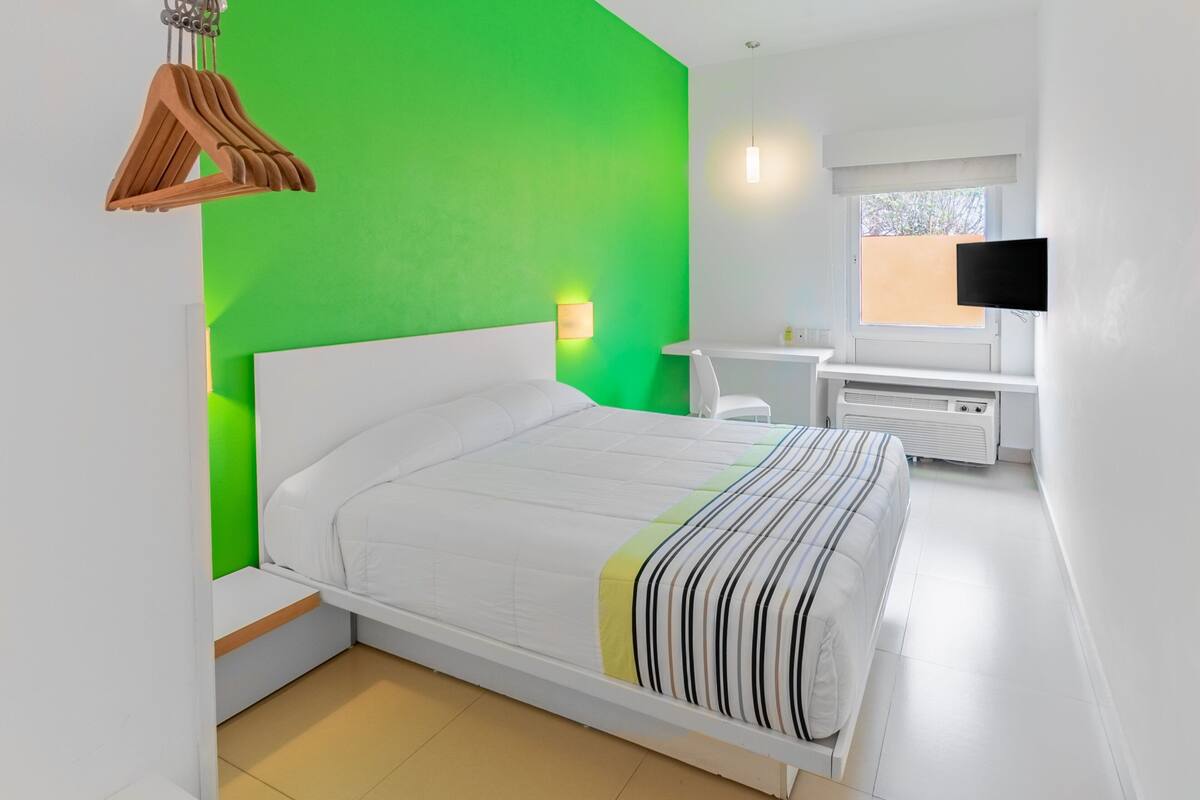 A modern bedroom with a green accent wall, a white bed, a bedside table, a chair, a TV, and a window.