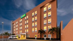 A multi-story hotel with a modern design, featuring a prominent green sign, multiple windows, and a parking area with a white van. - City Express Junior by Marriott Veracruz Aeropuerto (Veracruz)
