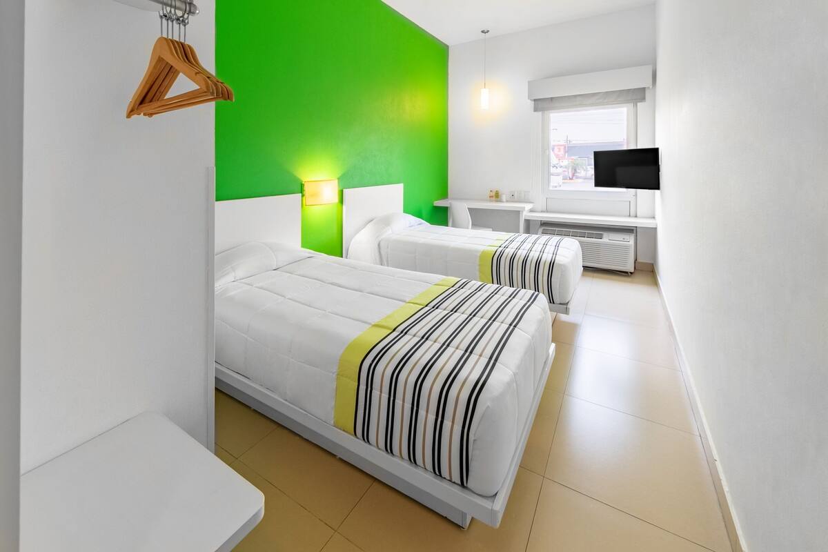 A hotel room with two beds, a green accent wall, a TV, and a window with a view.