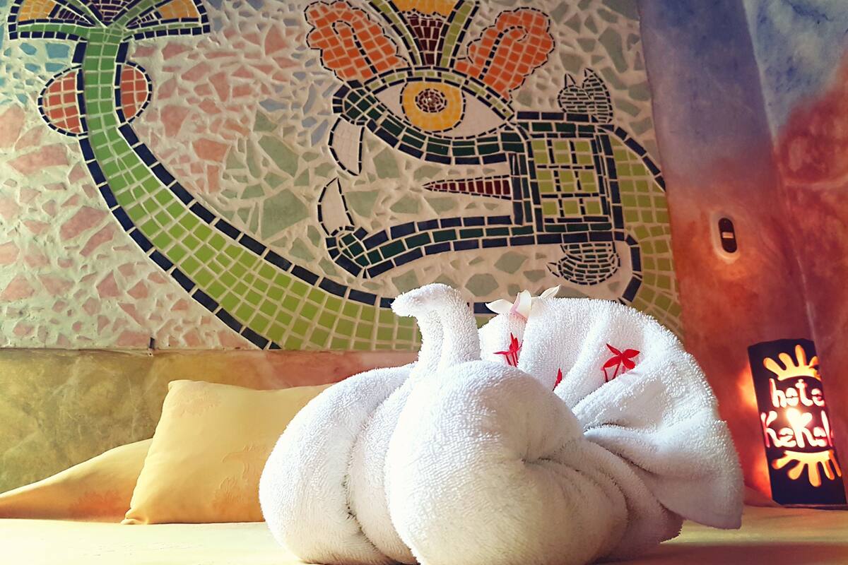 Un jouet en peluche en forme de tortue, posé sur un lit, avec un mur décoré de mosaïques en arrière-plan.
