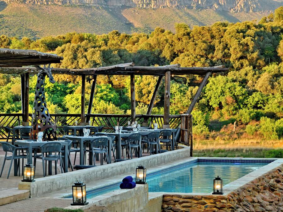 Un espacio de comedor al aire libre con mesas y sillas, una piscina y un paisaje montañoso de fondo.
