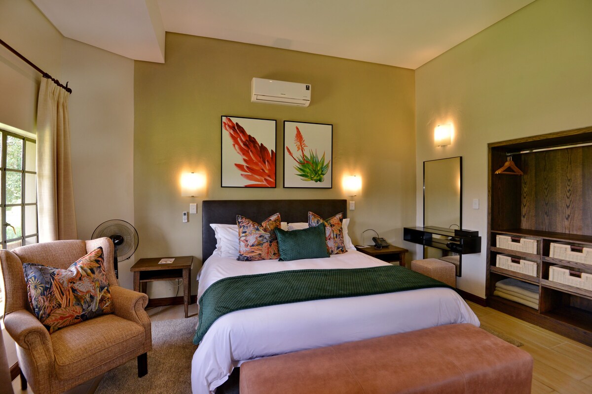 A hotel room with a bed, a chair, a desk, a fan, a mirror, and a wardrobe.