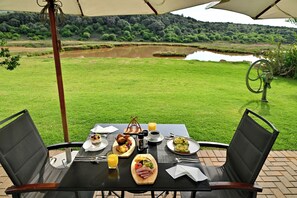 An outdoor dining table with chairs, a table setting with plates, cutlery, and a glass of orange juice, set against a backdrop of a grassy area and a body of water.