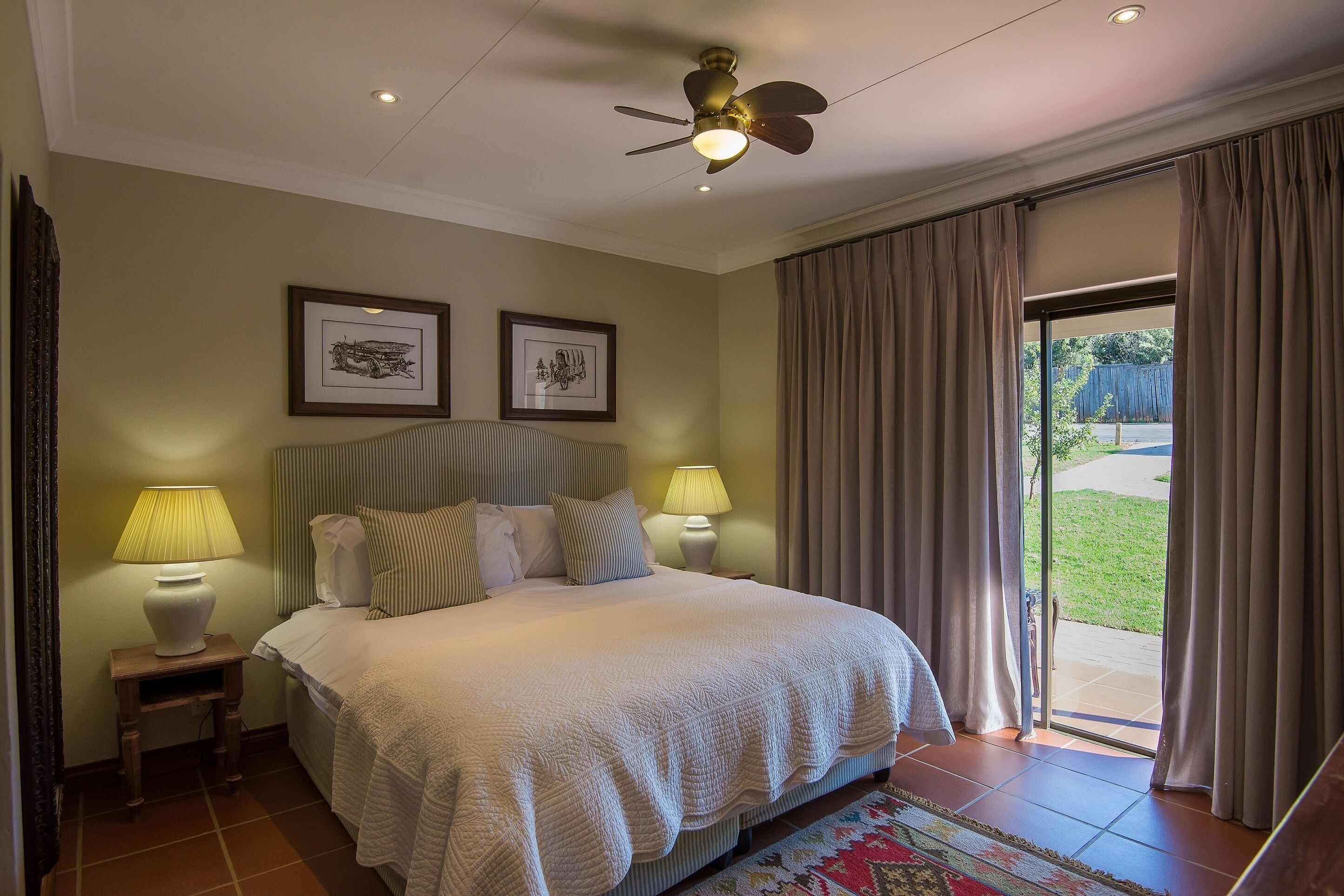 A bedroom with a bed, two bedside lamps, a ceiling fan, and a large window with curtains.
