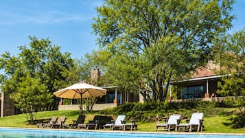 Piscina com espreguiçadeiras e guarda-sóis, cercada por árvores e um edifício.
