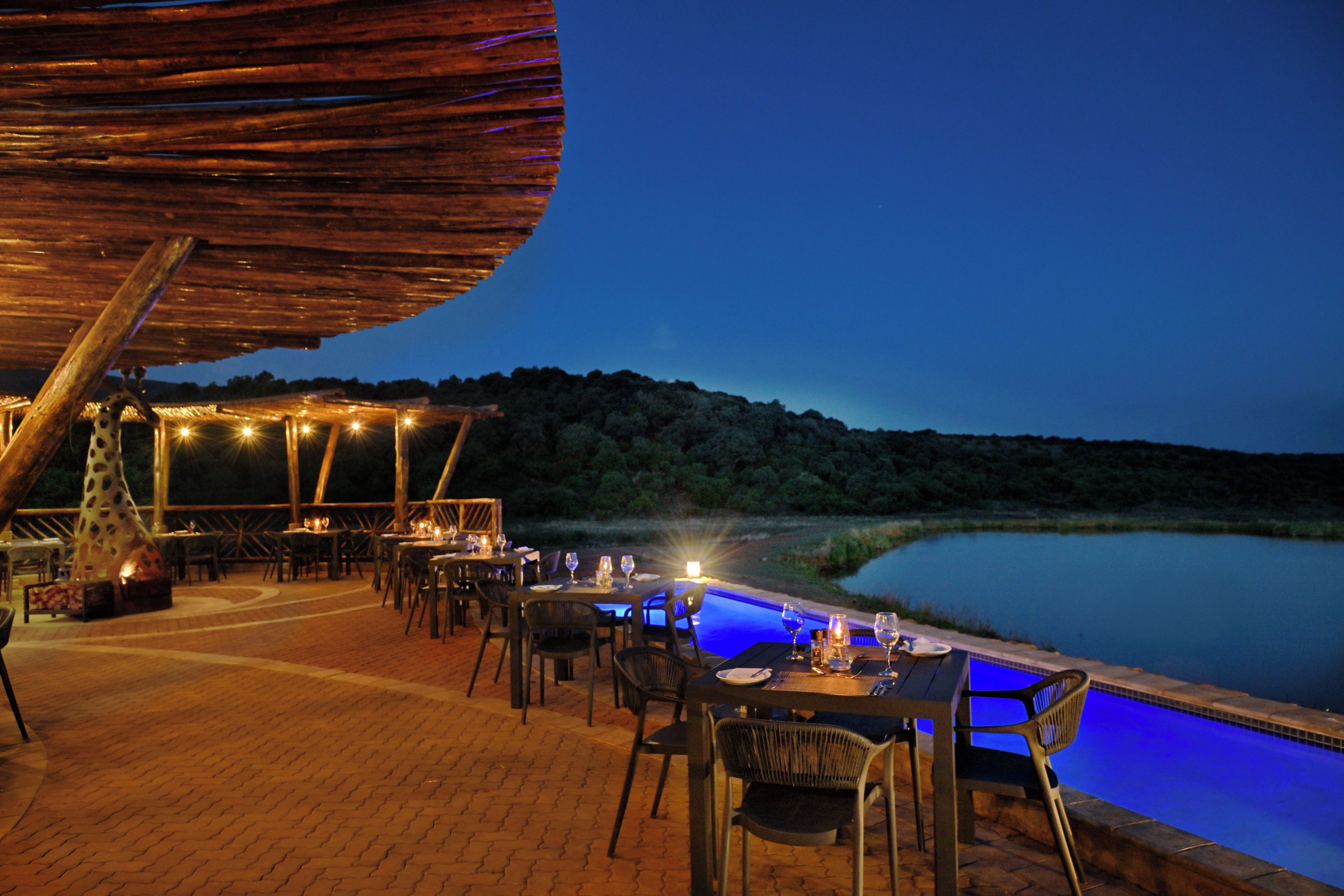 Un área de comedor al aire libre con mesas y sillas, piscina y vistas al agua y colinas.