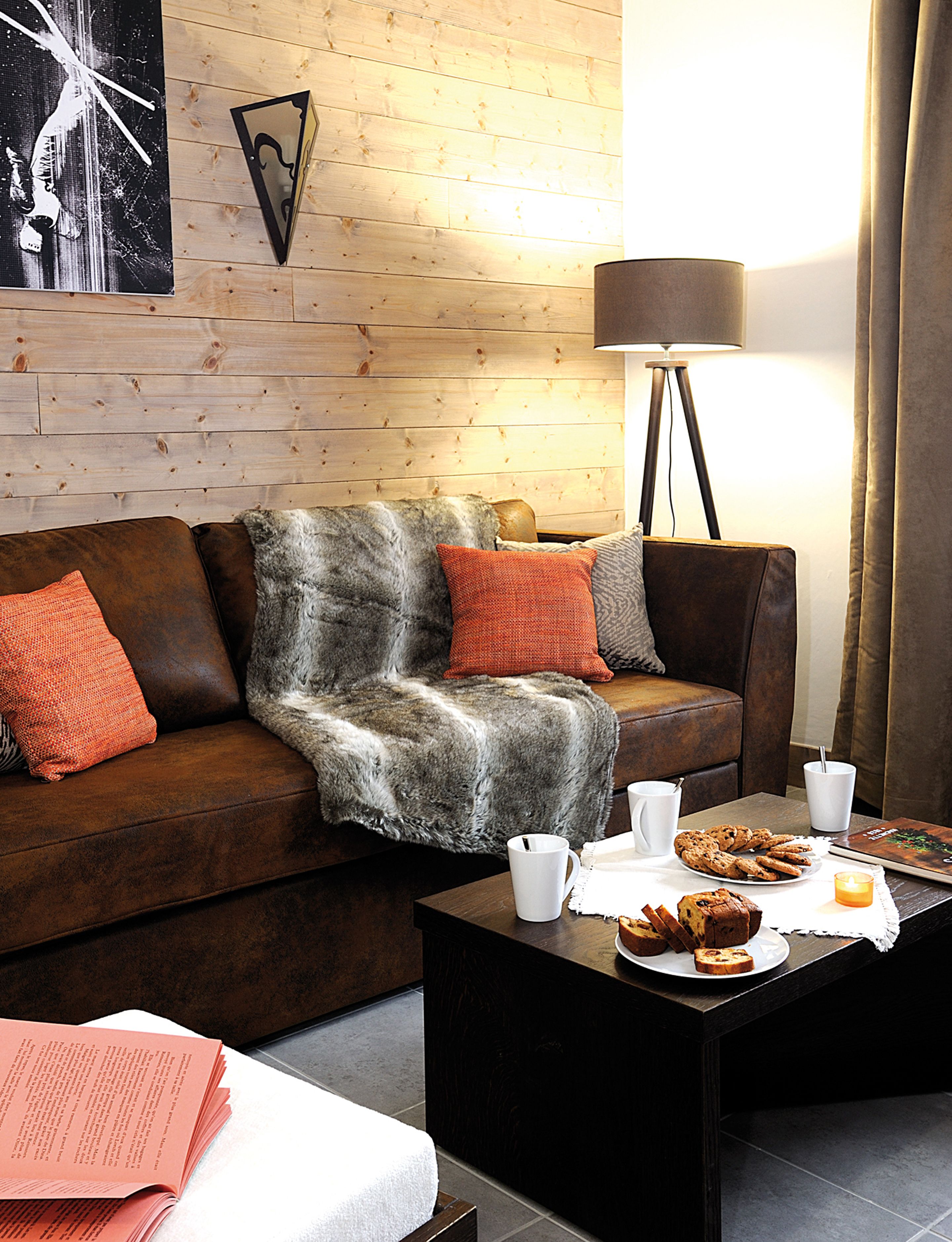 Un salon chaleureux avec un canapé en cuir marron, une couverture grise moelleuse et une table basse sur laquelle sont posés des en-cas et un livre.