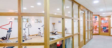 Un gimnasio con equipo de ejercicio, paredes de vidrio con estructura de madera y suelo de baldosas.