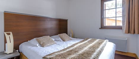 A bedroom with a wooden headboard, a large bed with white bedding, a fur throw, a bedside table with a lamp, and a window with curtains.