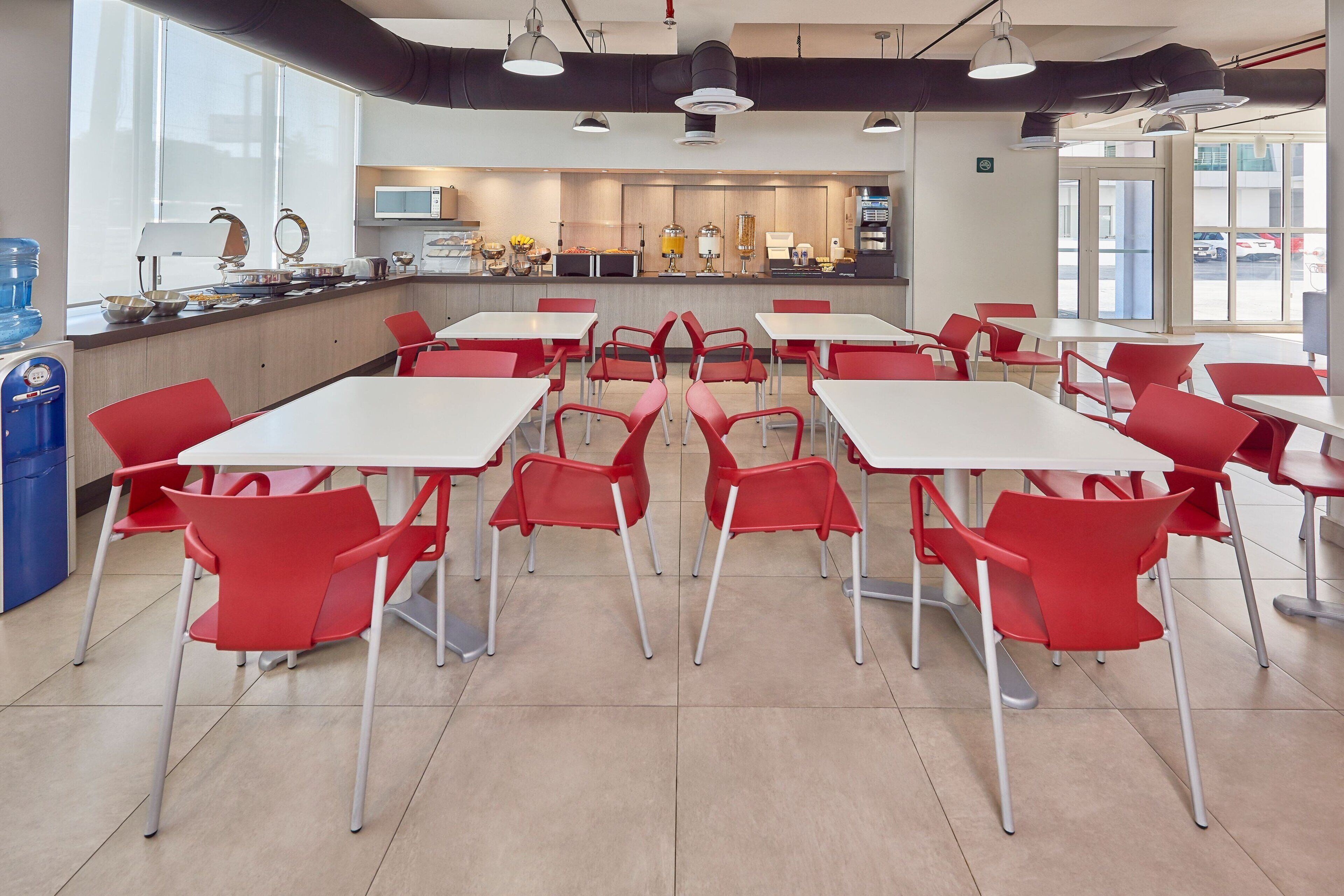 A modern cafeteria with red chairs and white tables, a kitchen area in the background, and large windows providing natural light.