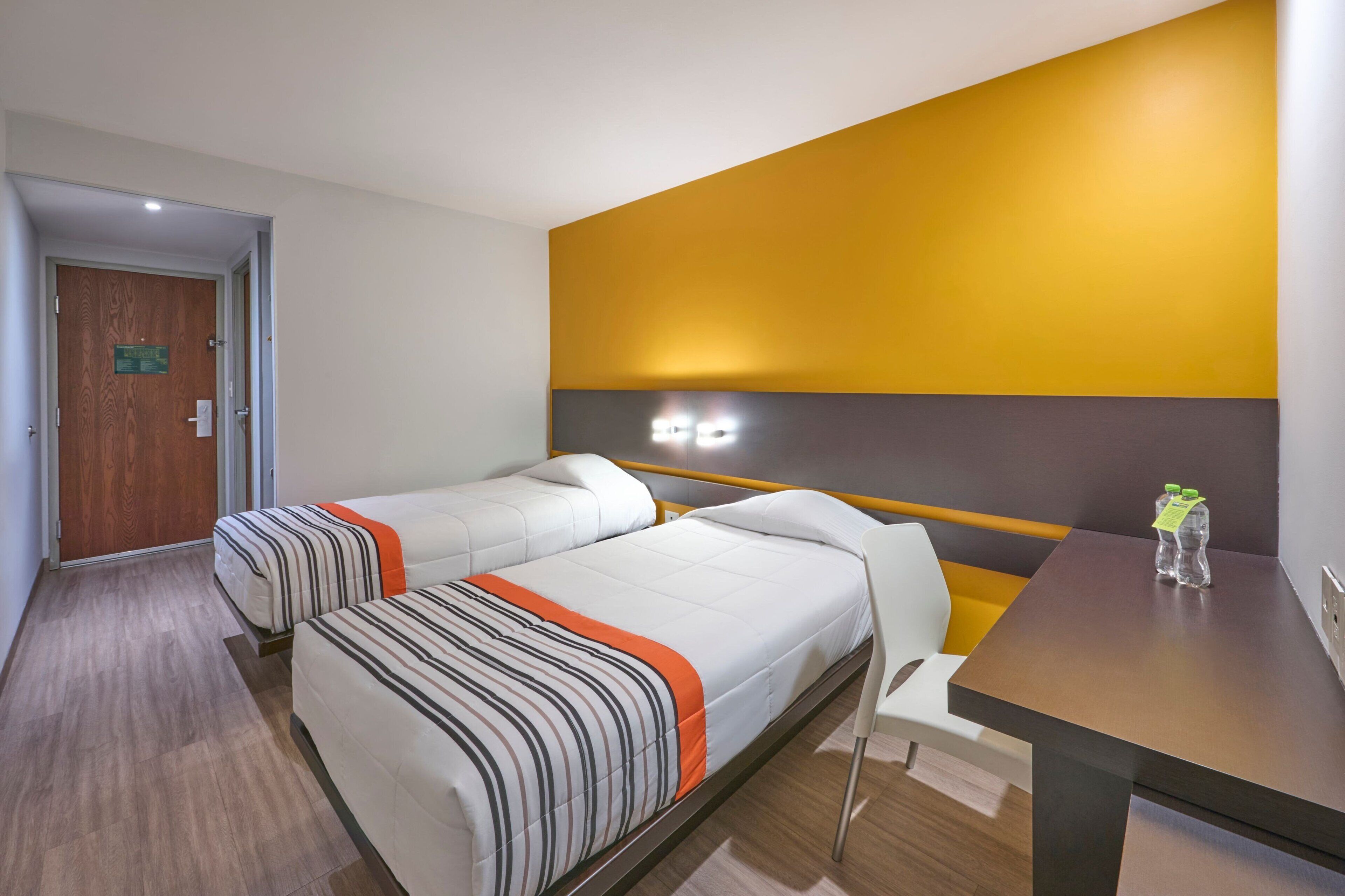 Ein Hotelzimmer mit zwei Betten, einer Holztür, einer gelben Akzentwand und einem Tisch mit einer Flasche.