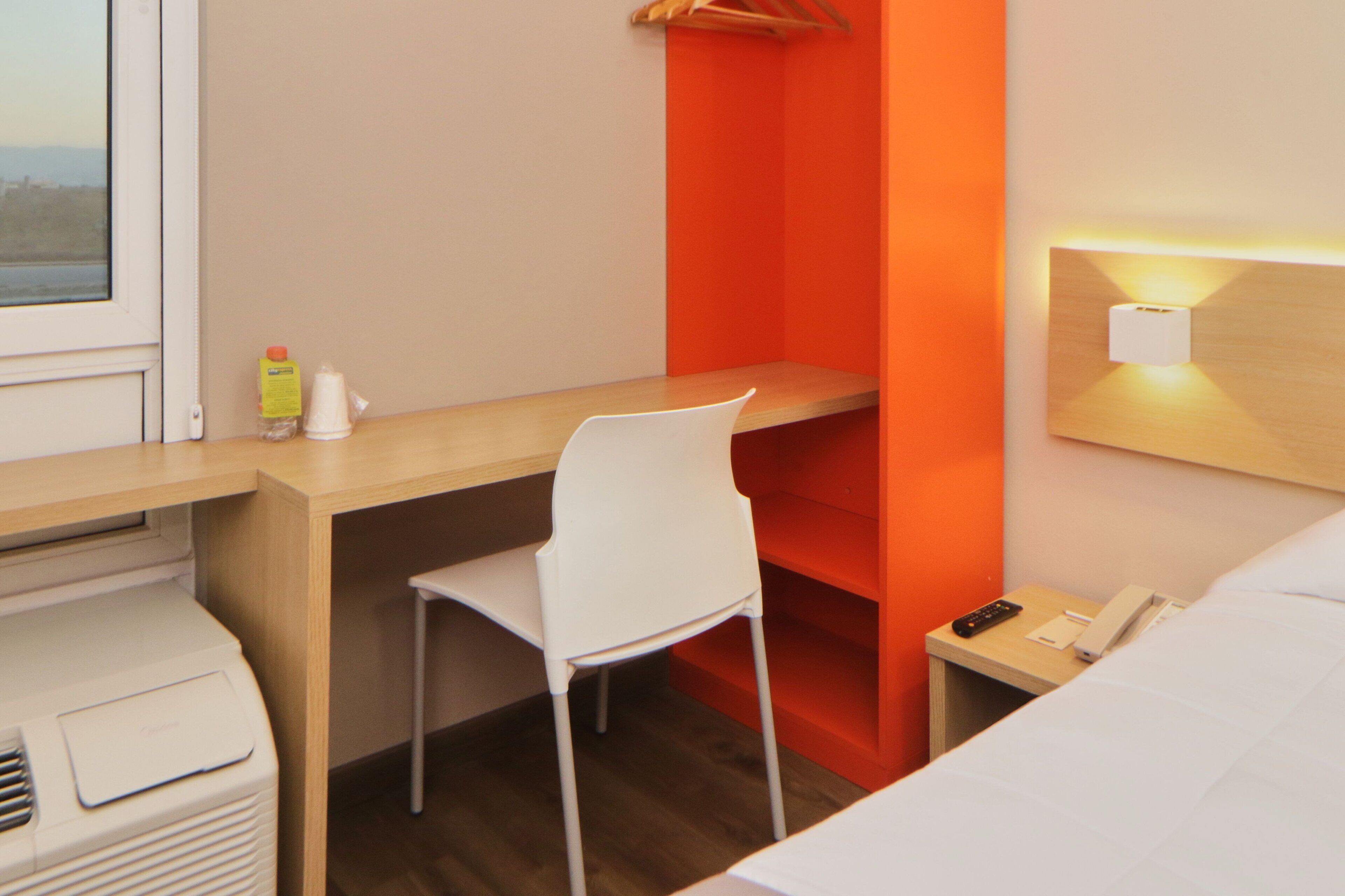 Een hotelkamer met een houten bureau, een witte stoel, een nachtkastje met telefoon en afstandsbediening, en een raam met uitzicht.