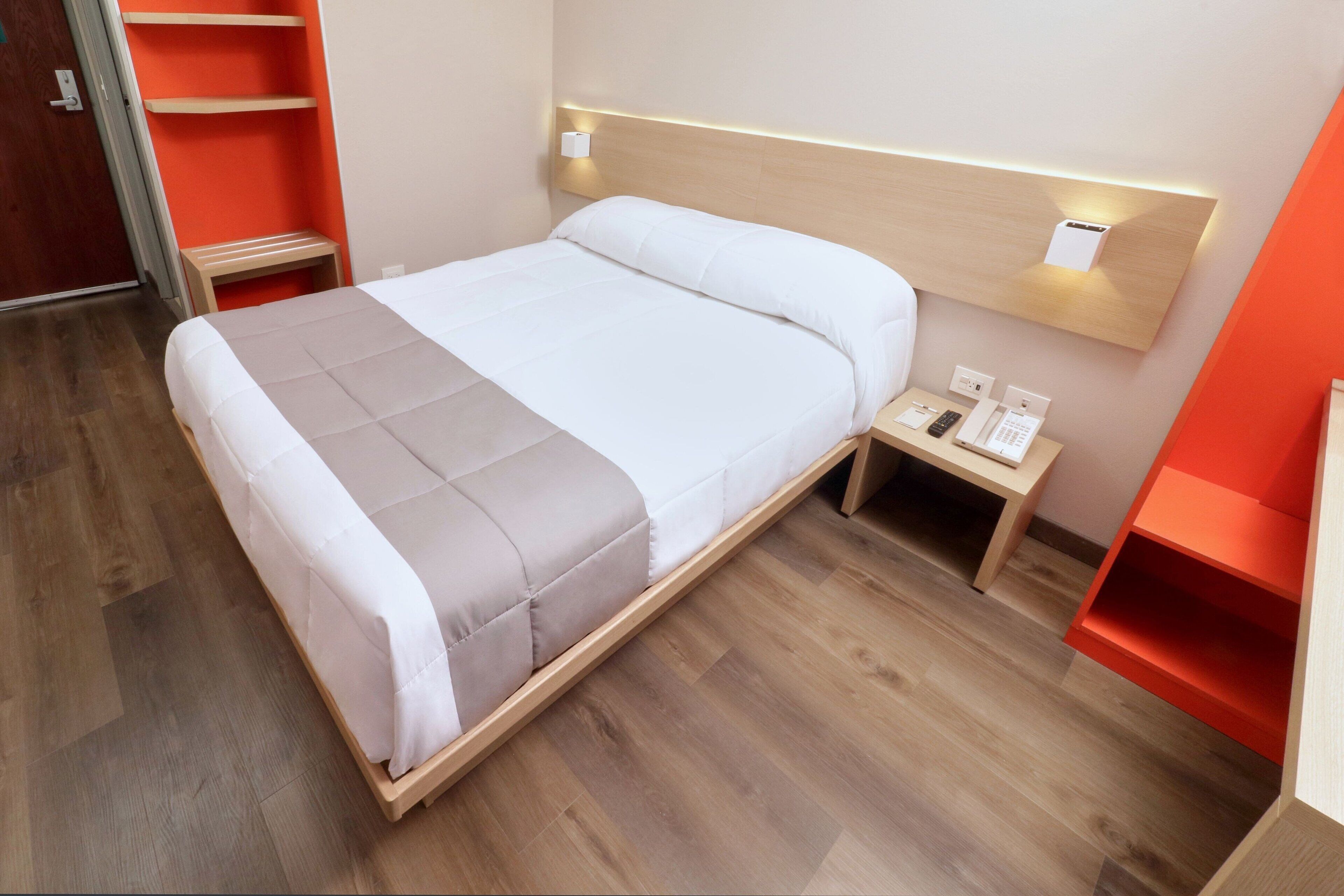 A neatly made bed with a wooden frame, a bedside table with a phone and a small lamp, and a red shelf unit to the right.