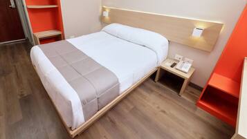 A neatly made bed with a wooden frame, a bedside table with a phone and a small lamp, and a red shelf unit to the right.