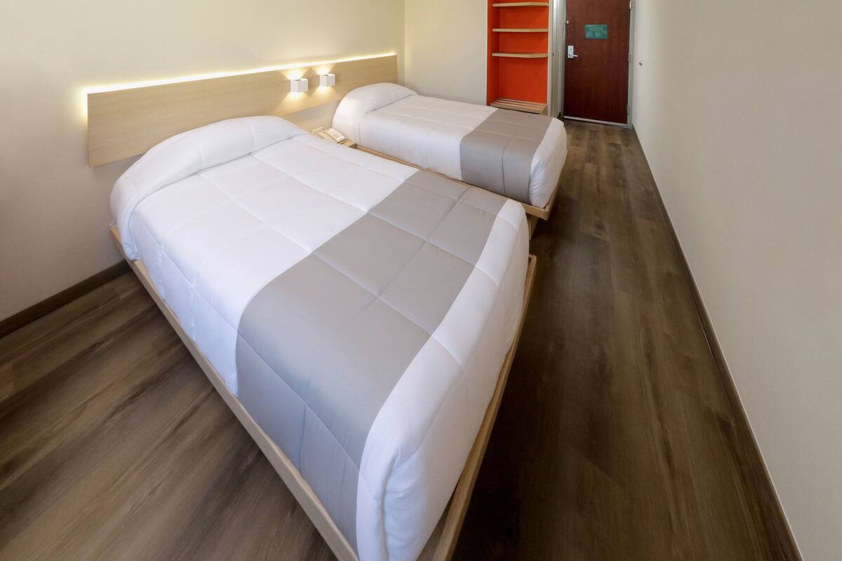 Une chambre d’hôtel avec deux lits, un plancher en bois, une table de chevet et un luminaire fixé au mur.