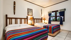 A hotel room with two beds, each with a colorful striped bedspread, a wooden headboard, bedside lamps, a decorative wall piece, and a window with curtains.