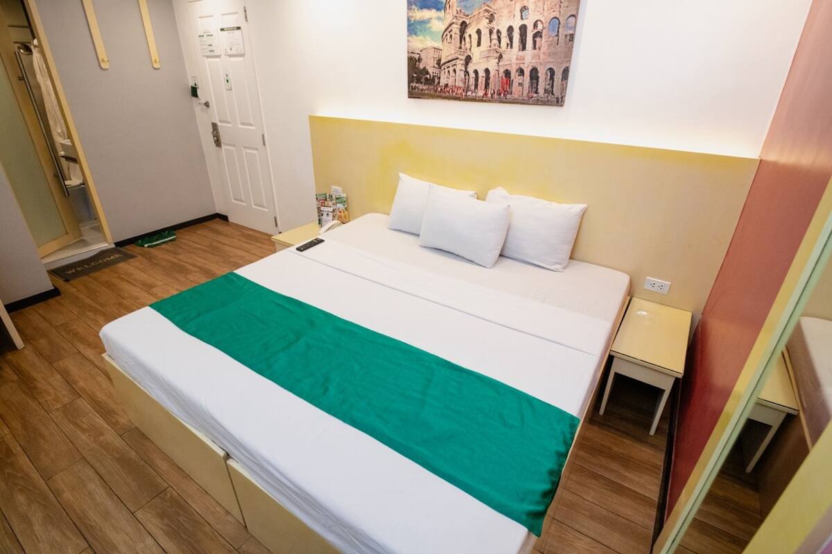 Une chambre d’hôtel avec un lit, une table de chevet, un tableau représentant un bâtiment historique et une porte.