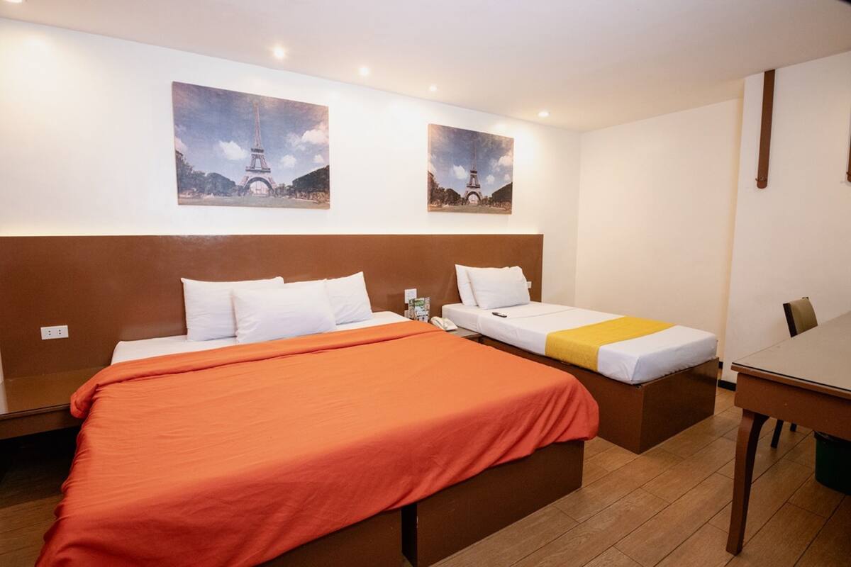 Une chambre d’hôtel avec un grand lit, un lit plus petit et un bureau. Deux tableaux représentant la Tour Eiffel sont accrochés au mur.