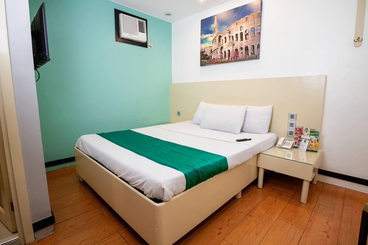 Une chambre d’hôtel avec un lit, une table de chevet avec un téléphone et des brochures, un tableau au mur et un appareil de climatisation.