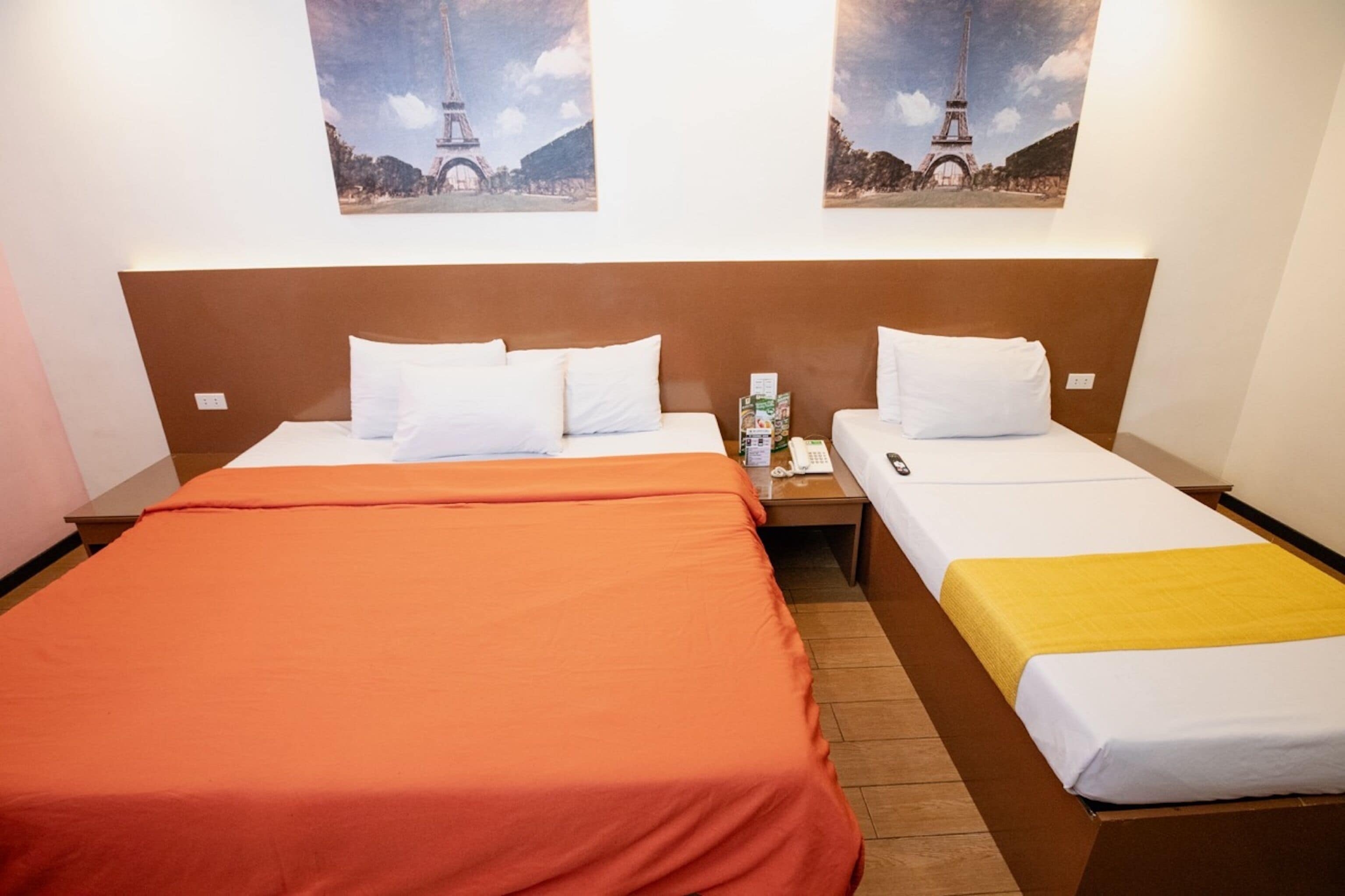 A hotel room with two beds, one with an orange bedspread and the other with a yellow one. There are two nightstands with a phone and a bottle on each. The room has a wooden headboard and two framed pictures of the Eiffel Tower on the wall.