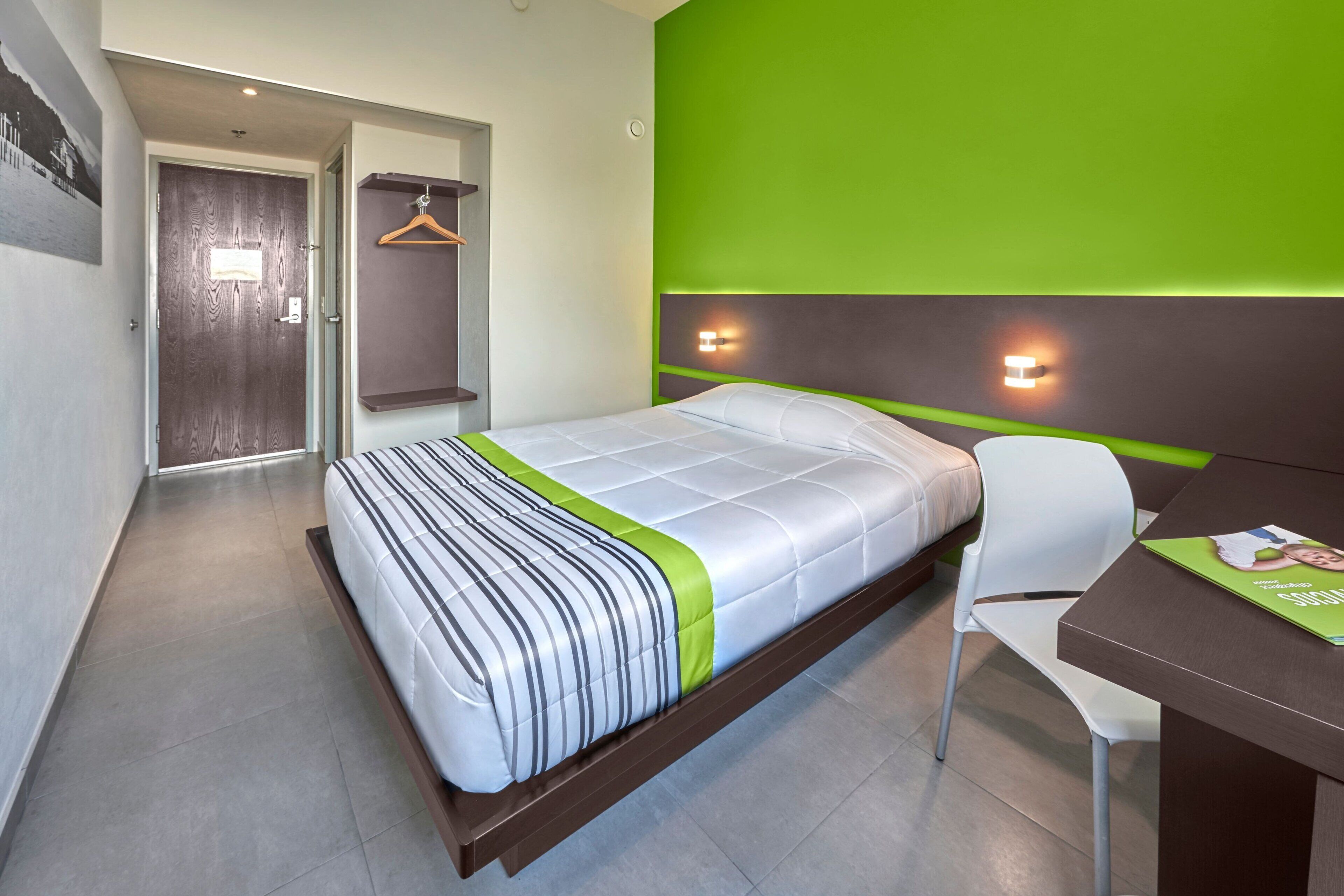 Una habitación de hotel moderna con una cama, un escritorio y una silla. Cuenta con una pared verde de acento y una puerta con interruptor de luz.