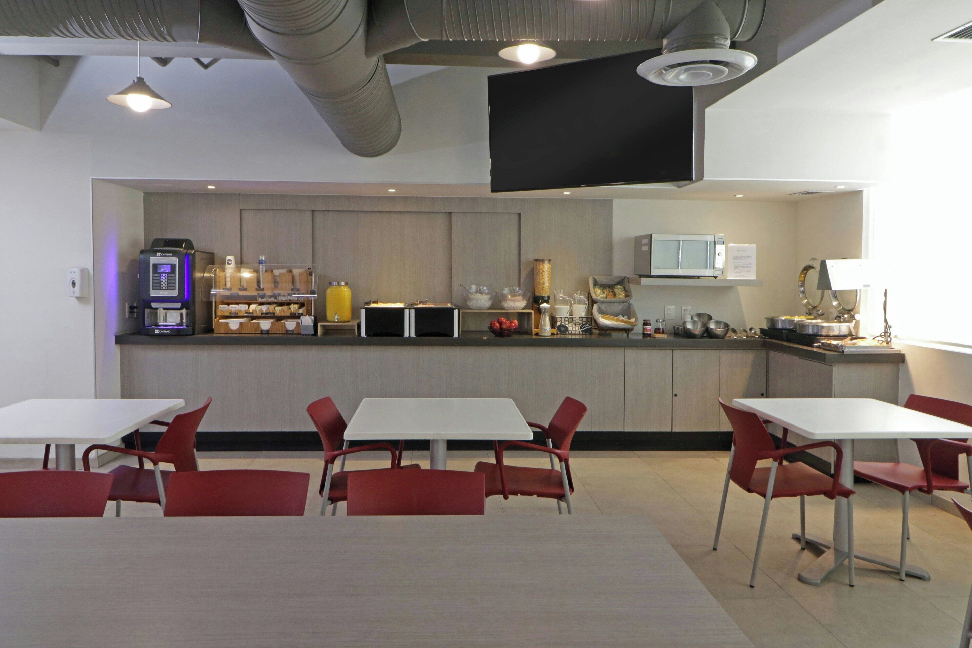 A modern kitchen with white cabinets, a microwave, and a coffee machine. There are red chairs and a white table in the dining area.