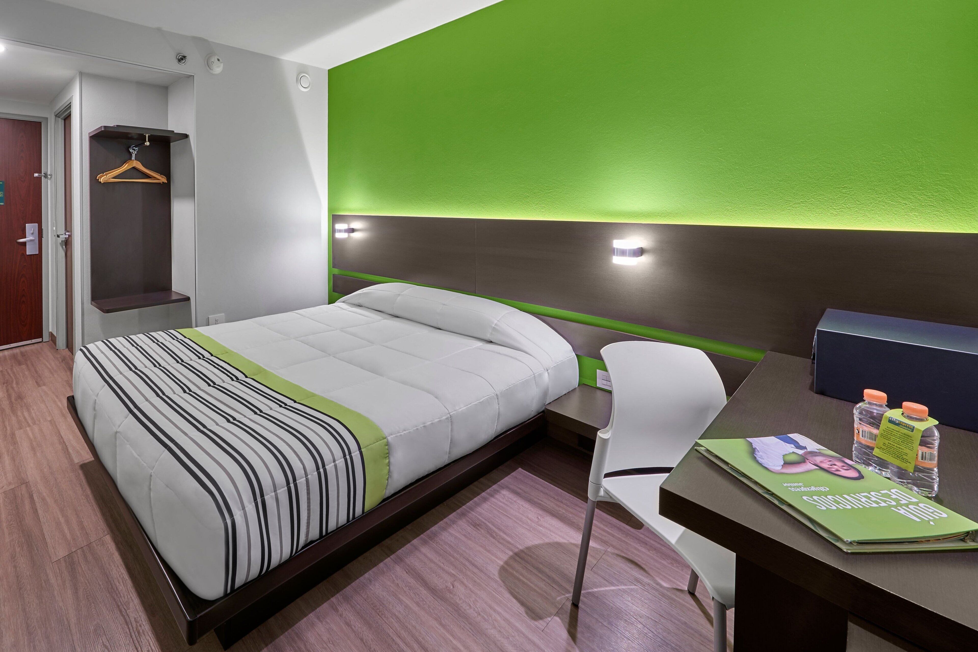 A hotel room with a bed, a desk, a chair, a hanging coat rack, and a green wall.
