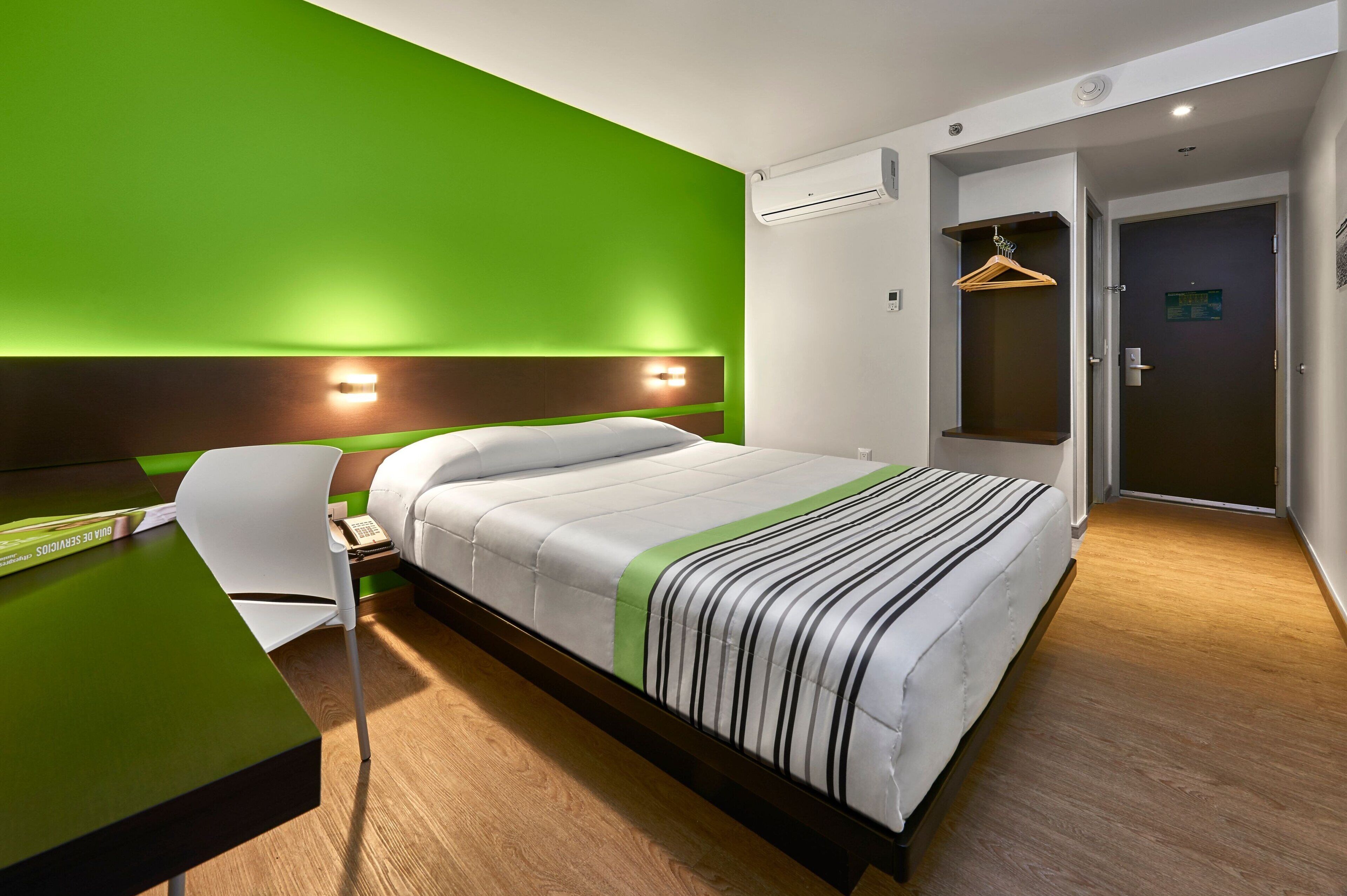 Quarto de hotel moderno com parede verde de destaque, cama com colcha listrada, escrivaninha com cadeira e um cabideiro suspenso.