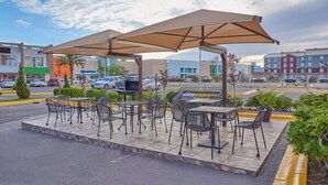 Een terras buiten met tafels en stoelen, een grote parasol, en zicht op een parkeerplaats en gebouwen op de achtergrond.