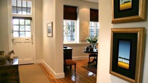 A hallway with wooden flooring, a door, and two windows with blinds. There are framed pictures on the wall and a small table with a vase. - Four Rosmead (Cape Town)