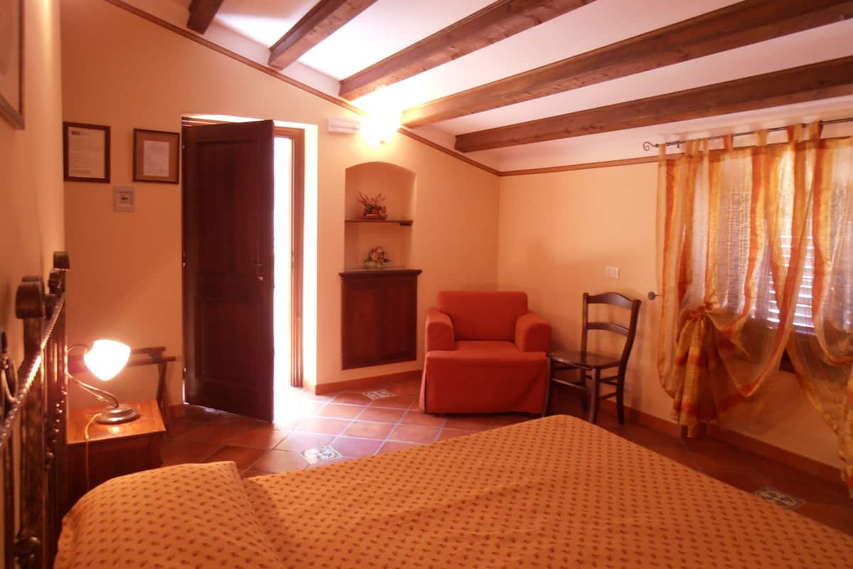 A room with a bed, a chair, a small table, a shelf with decorative items, and a window with curtains.