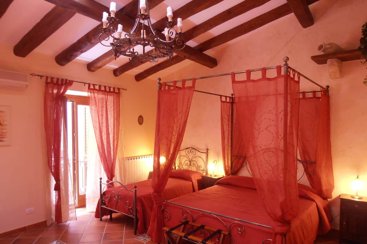 A four-poster canopy bed with a wooden frame and red bedding, a chandelier, and a tiled floor.