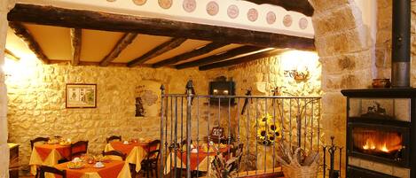 A rustic dining area with stone walls, wooden beams, and a fireplace. Tables are set with red tablecloths and chairs.