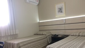A hotel room with two single beds, a desk, a chair, an air conditioning unit, a framed picture on the wall, and a bedside table with a phone and book. - Barigui Park Hotel (Curitiba)