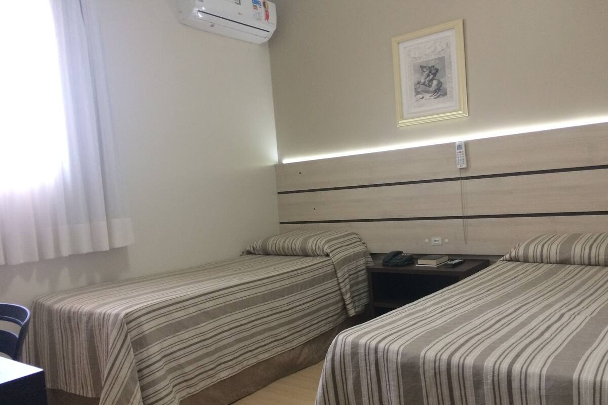 A hotel room with two single beds, a desk, a chair, an air conditioning unit, a framed picture on the wall, and a bedside table with a phone and book.