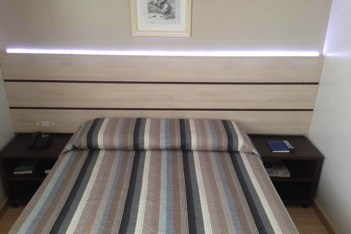 A neatly made bed with striped bedding, a headboard with built-in lighting, and bedside tables with a remote control and a book.