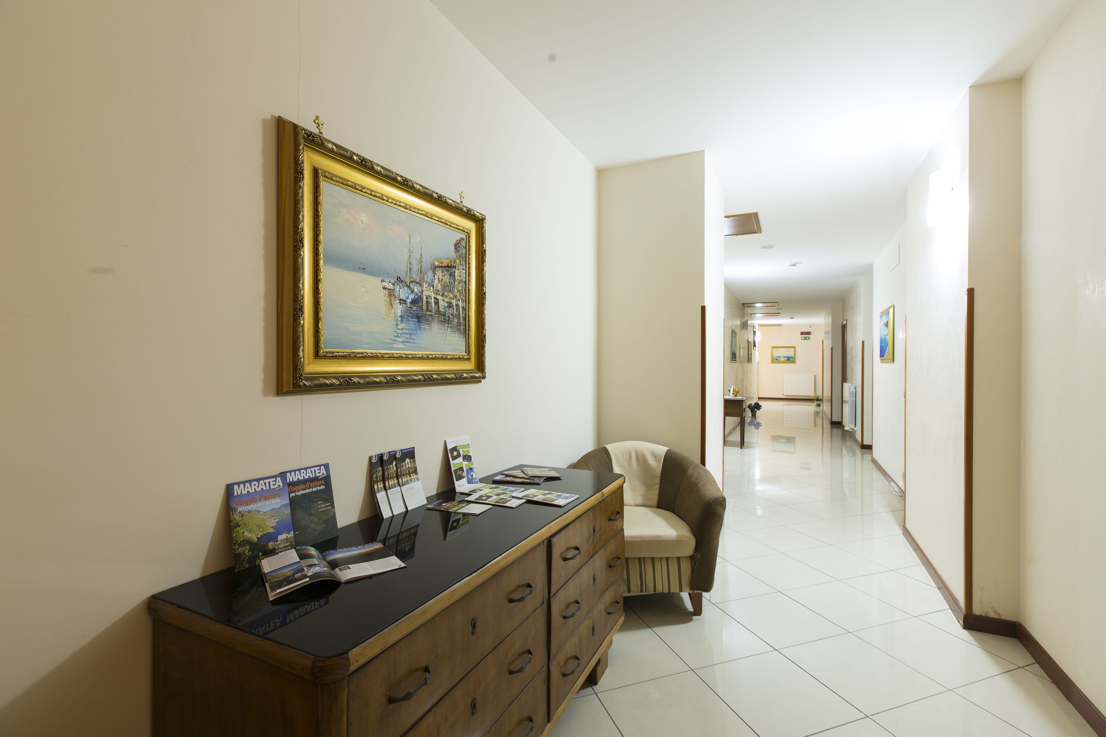 A hotel reception area with a wooden desk, brochures, a framed painting, and a chair.