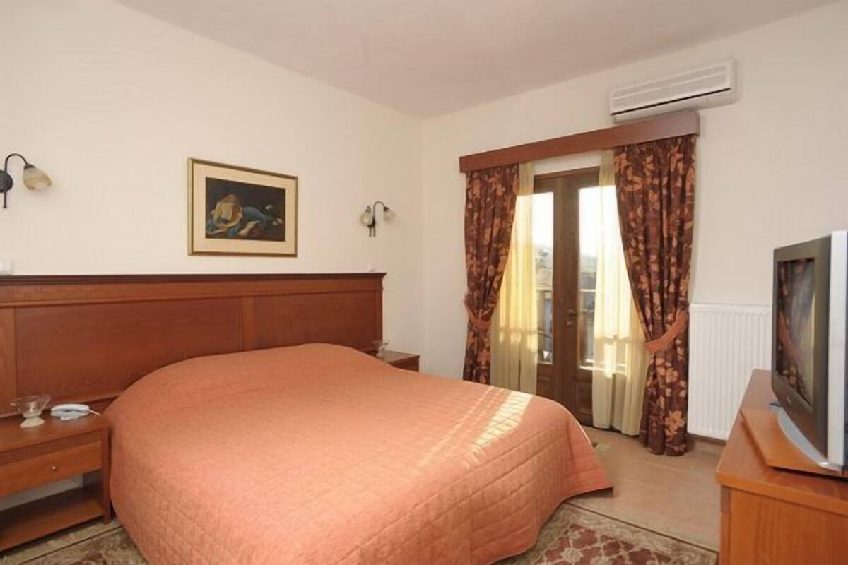 Ein Hotelzimmer mit einem großen Bett, einem Holz-Kopfteil, einem Bild an der Wand, einem Fernseher auf einem Holztisch und einem Fenster mit Vorhängen.