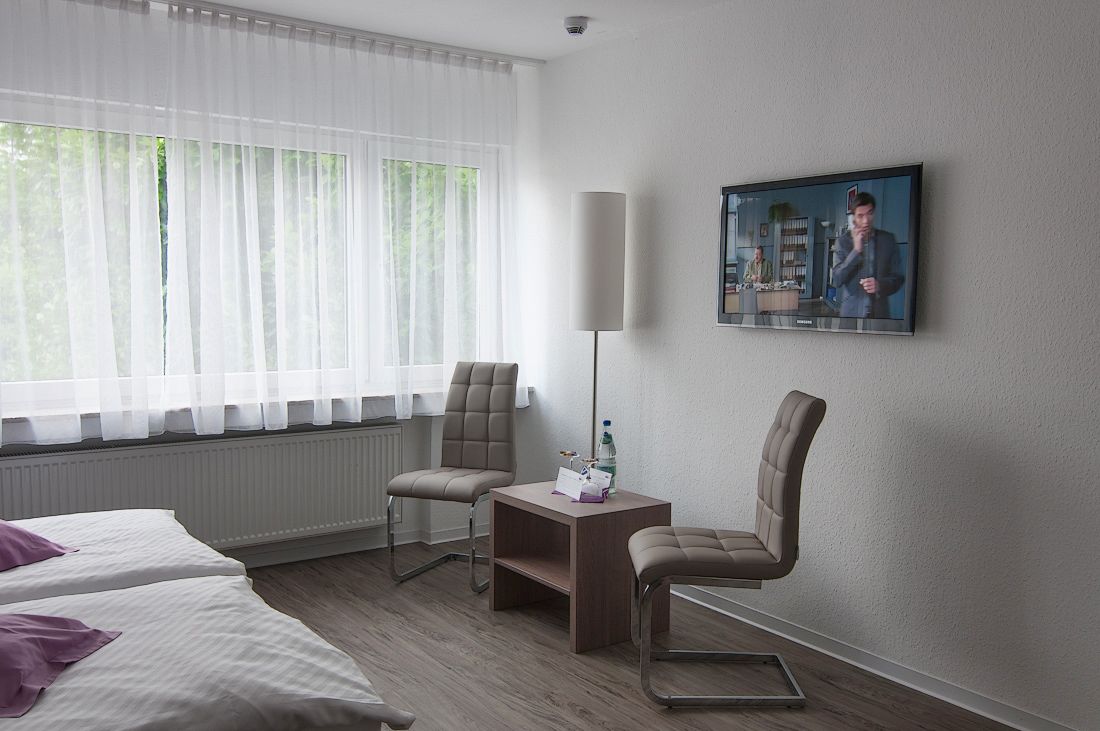 A hotel room with two beds, a desk, a chair, a standing lamp, and a TV on the wall.