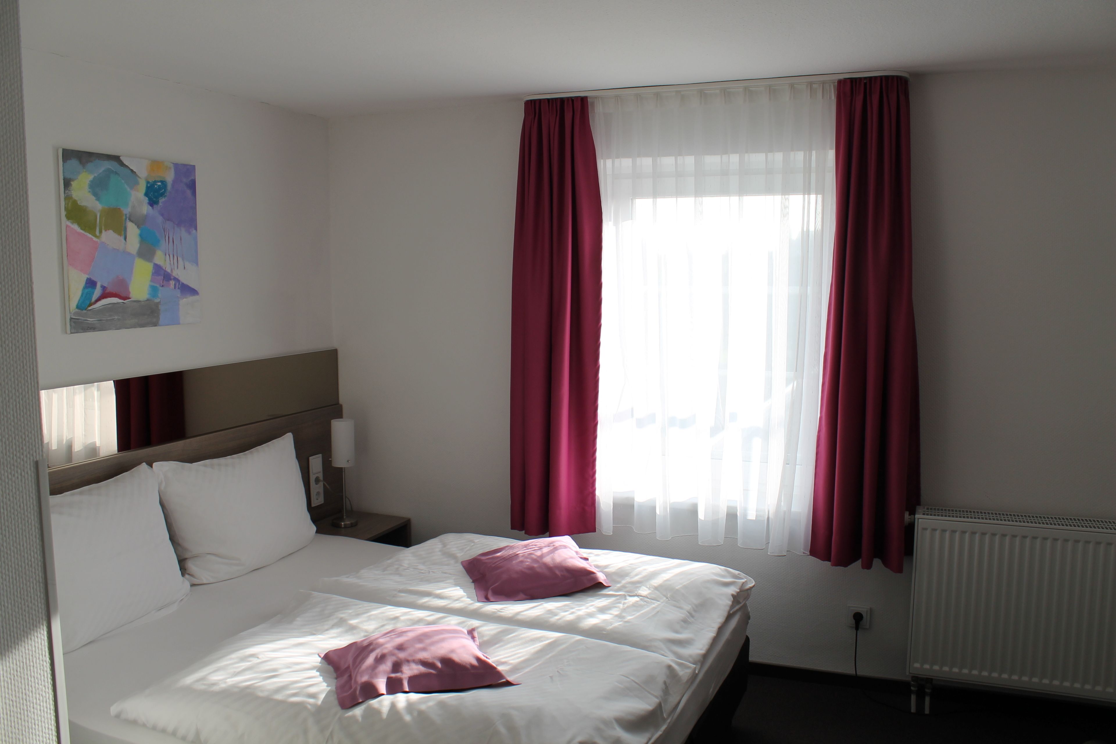 A hotel room with two beds, white bedding, a painting on the wall, and a window with red curtains.
