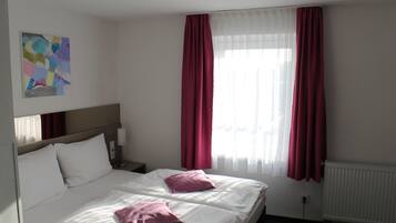 A hotel room with two beds, white bedding, a painting on the wall, and a window with red curtains.