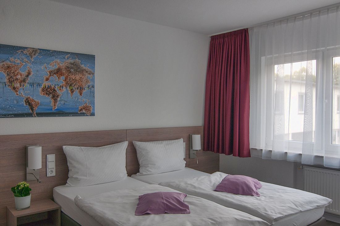 A hotel room with two beds, a world map on the wall, a window with curtains, and a small plant on the nightstand.