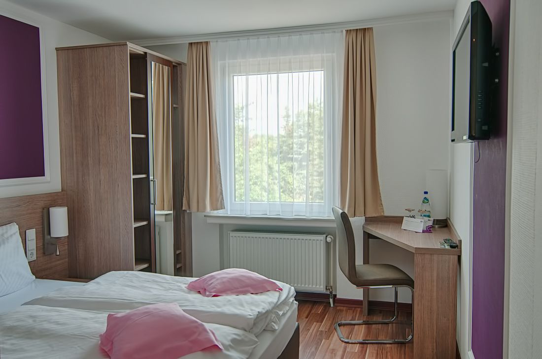 A hotel room with a bed, a desk, a chair, a TV, and a window with curtains.