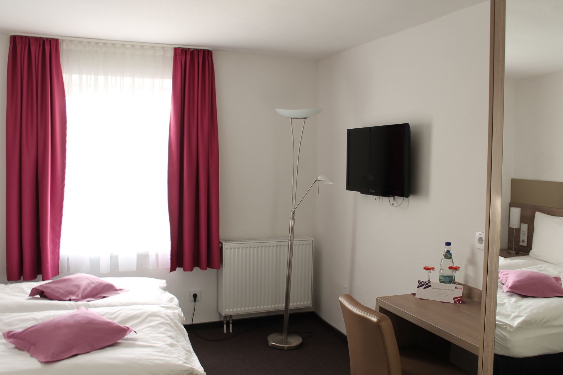 A hotel room with two beds, a desk, a chair, a floor lamp, a TV, and a radiator.