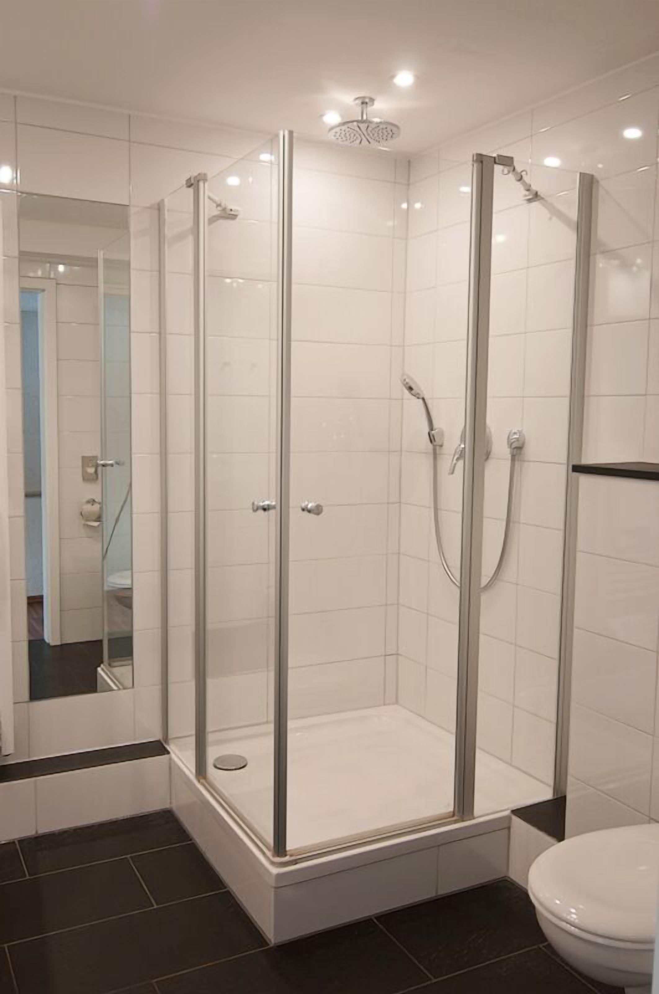 Un baño moderno con cabina de ducha de vidrio, paredes con azulejos blancos y suelo de baldosas negras.