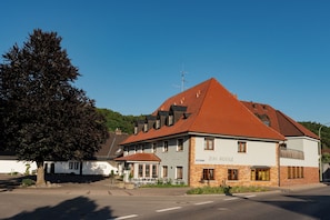 Um edifício de dois andares com telhado de telhas vermelhas, uma placa com a inscrição 'Zum Rossle' e uma grande árvore em frente.