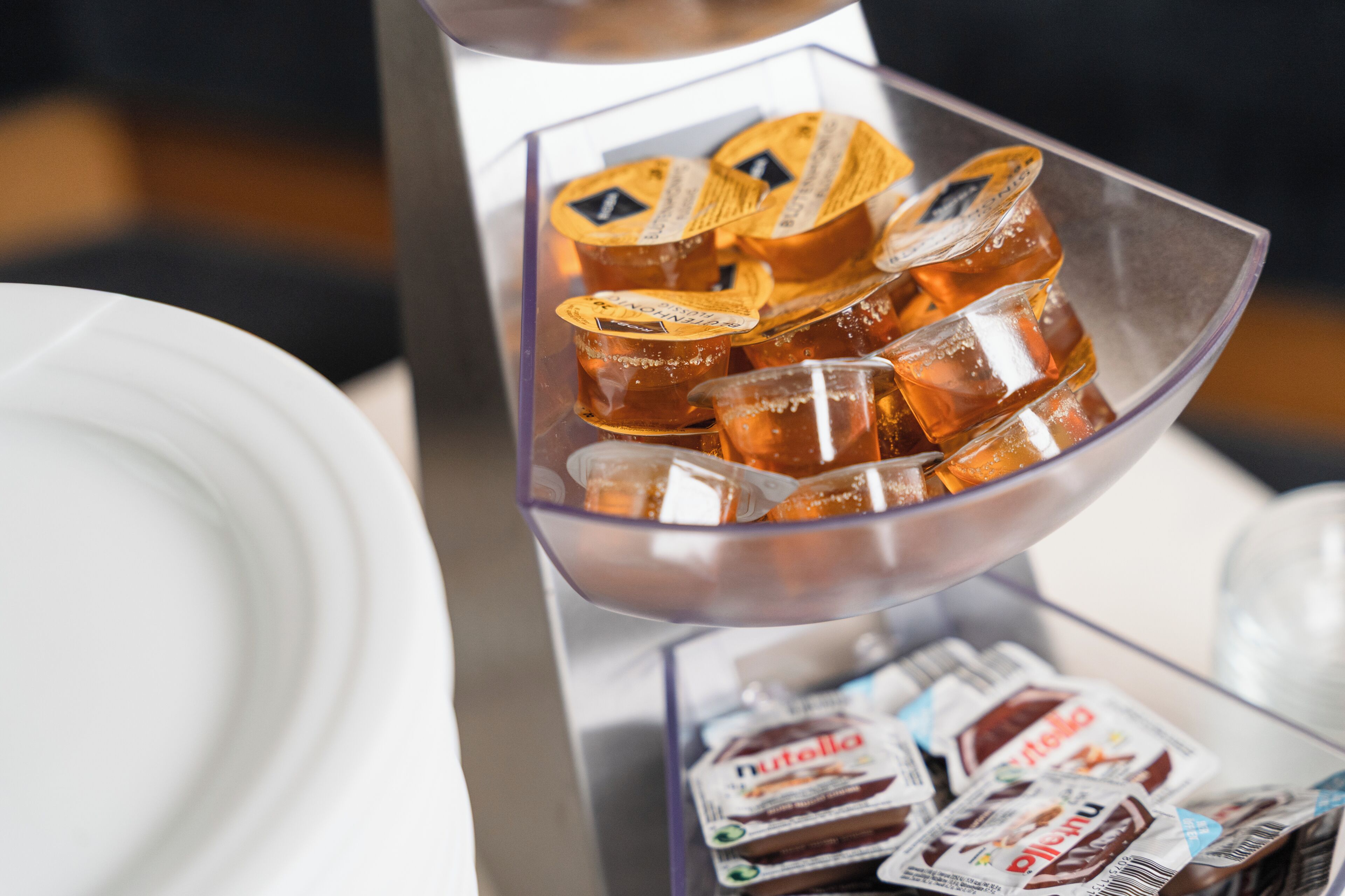 En klar glasbeholder med flere små, individuelt indpakkede genstande, sandsynligvis mad eller snacks, samt en hvid tallerken og et glas i forgrunden.